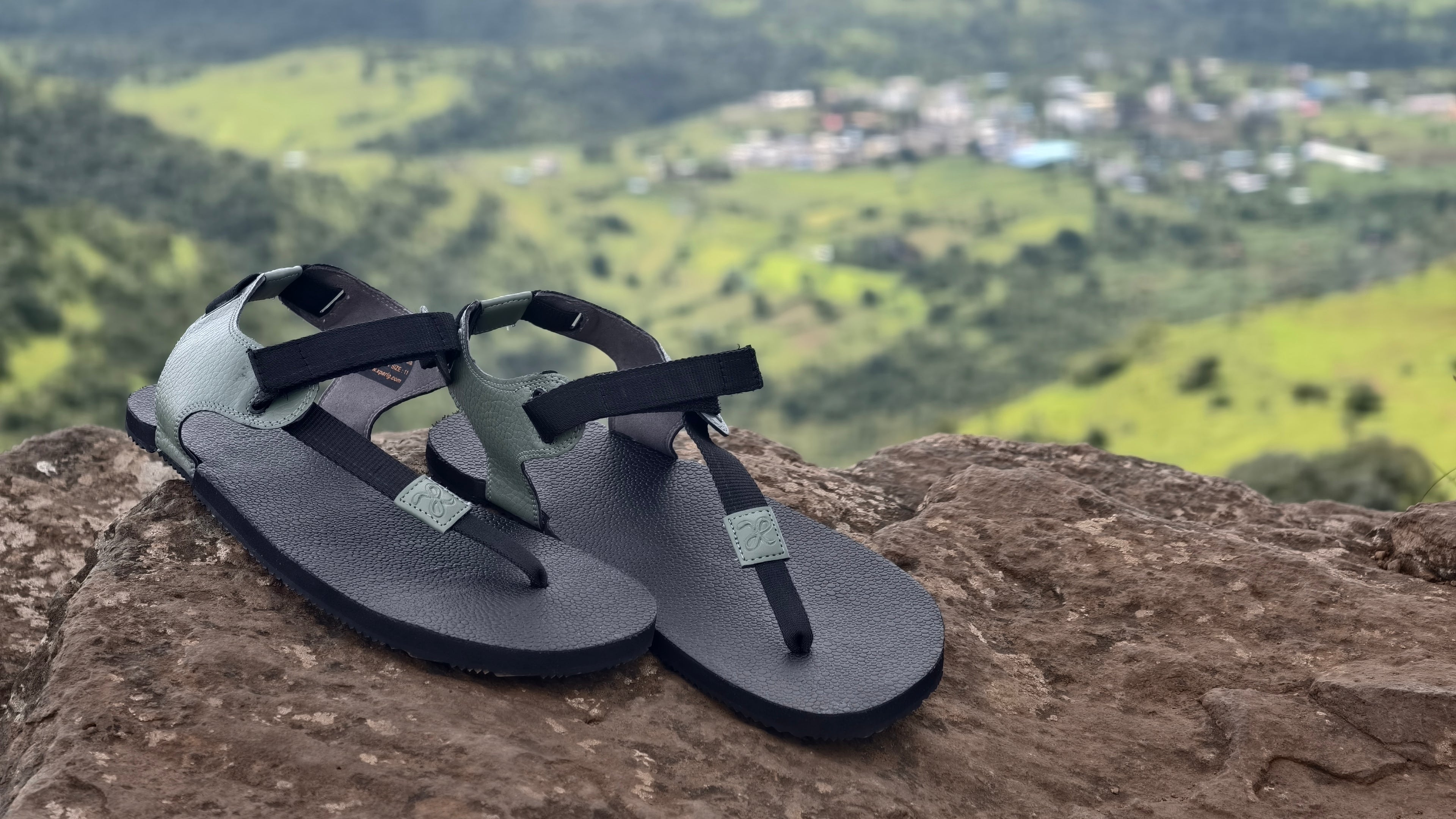 Black sandals on a rock with a scenic background of green hills and buildings.